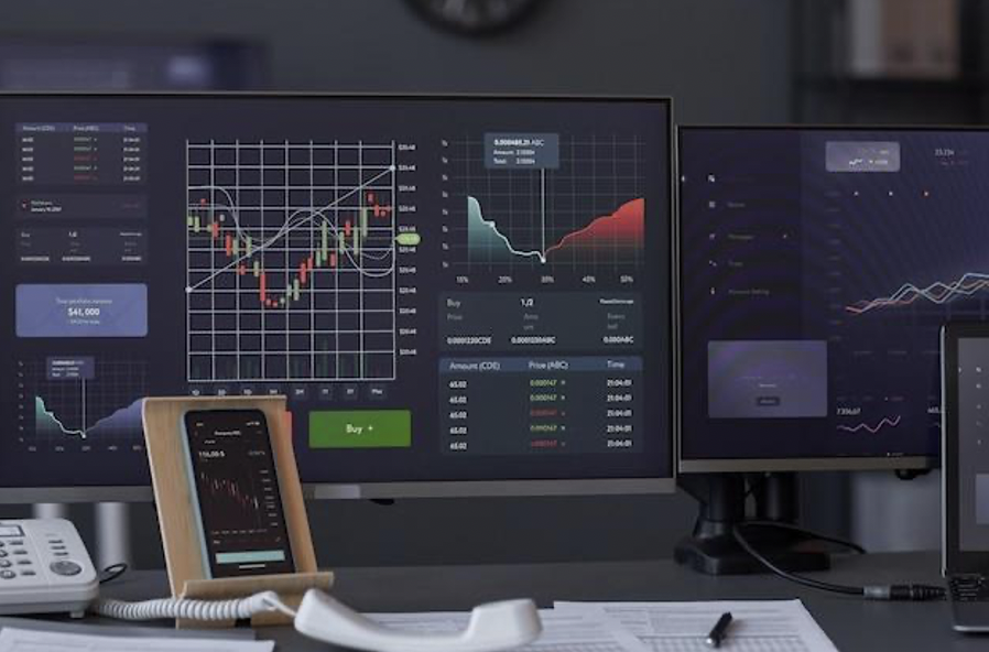 A trading desk setup with multiple monitors displaying financial graphs and a smartphone placed on the desk